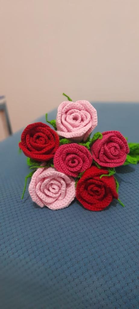 Close-up of handmade crochet roses in a variety of colors, including red, yellow, purple, and pink. The petals are intricately crocheted and feature a soft texture. The center of each rose is a contrasting color and has a raised, textured appearance.