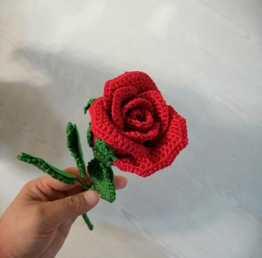 Close-up of handmade crochet roses in a variety of colors, including red, yellow, purple, and pink. The petals are intricately crocheted and feature a soft texture. The center of each rose is a contrasting color and has a raised, textured appearance.