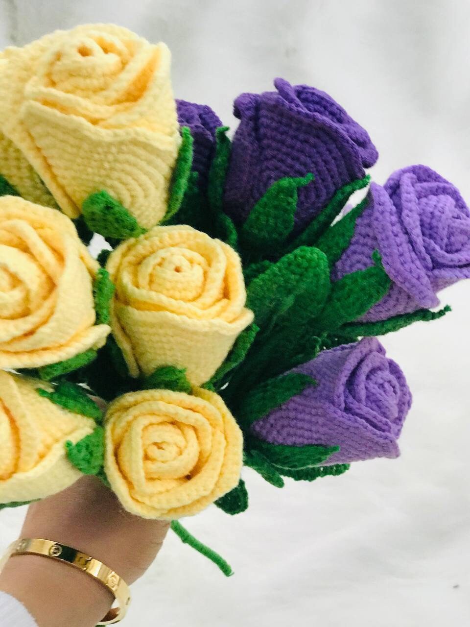 Close-up of handmade crochet roses in a variety of colors, including red, yellow, purple, and pink. The petals are intricately crocheted and feature a soft texture. The center of each rose is a contrasting color and has a raised, textured appearance.