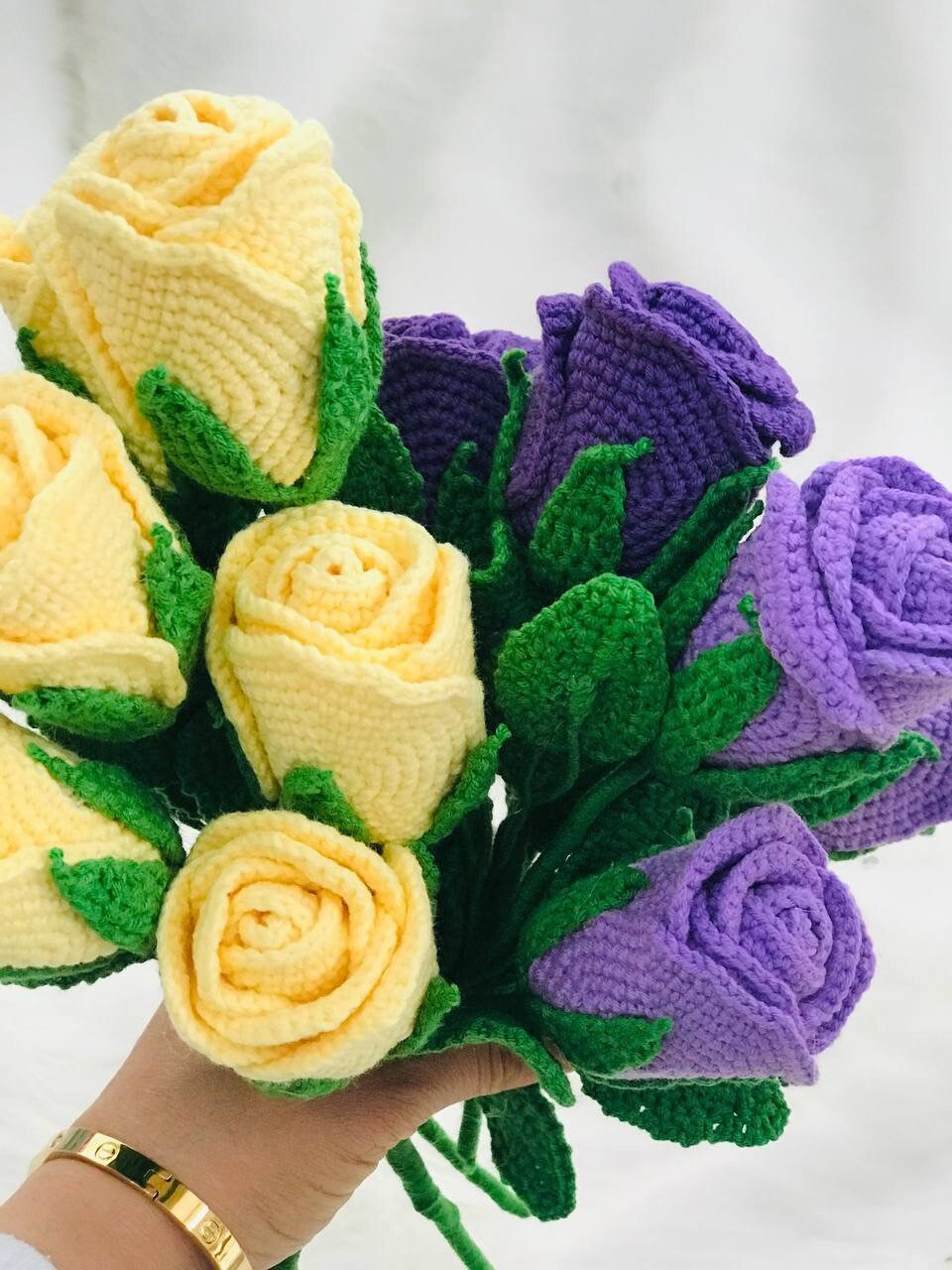 Close-up of handmade crochet roses in a variety of colors, including red, yellow, purple, and pink. The petals are intricately crocheted and feature a soft texture. The center of each rose is a contrasting color and has a raised, textured appearance.