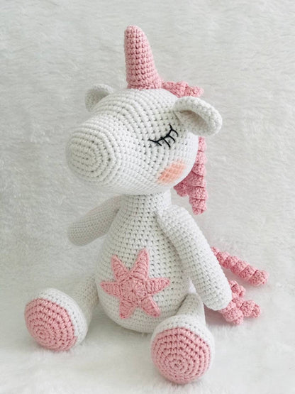 A handmade crochet unicorn plushie in pink and purple, featuring a unique design with a rainbow colored mane and tail. perfect for nursery decor or playtime, and soft and cuddly to the touch. The unicorn doll also has a sparkly horn and pastel colors