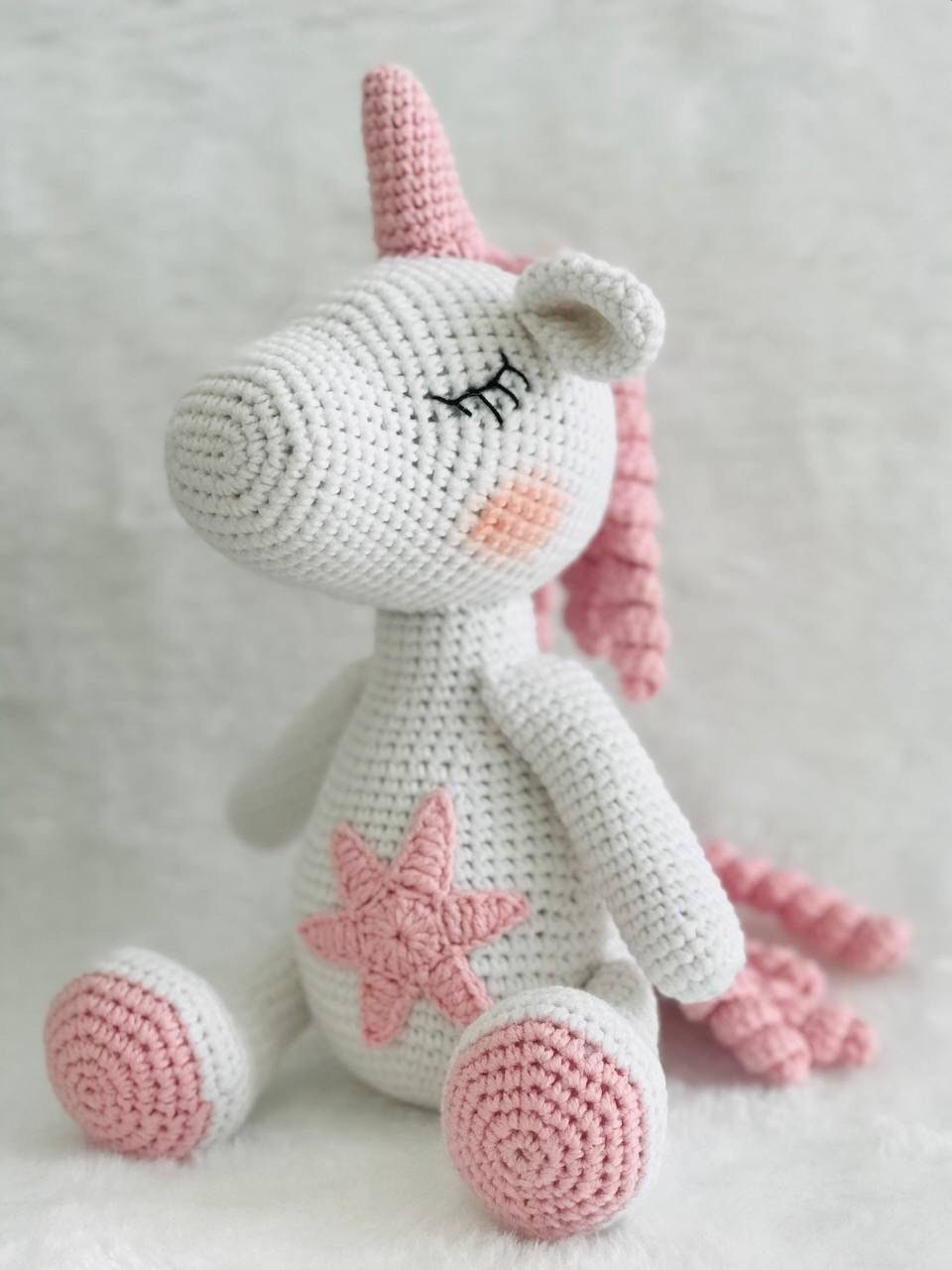 A handmade crochet unicorn plushie in pink and purple, featuring a unique design with a rainbow colored mane and tail. perfect for nursery decor or playtime, and soft and cuddly to the touch. The unicorn doll also has a sparkly horn and pastel colors