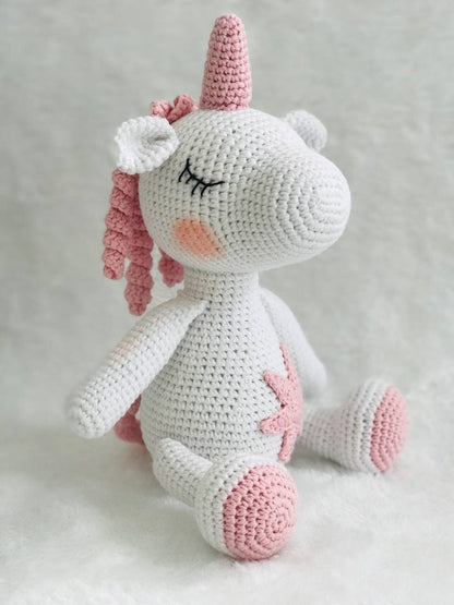 A handmade crochet unicorn plushie in pink and purple, featuring a unique design with a rainbow colored mane and tail. perfect for nursery decor or playtime, and soft and cuddly to the touch. The unicorn doll also has a sparkly horn and pastel colors