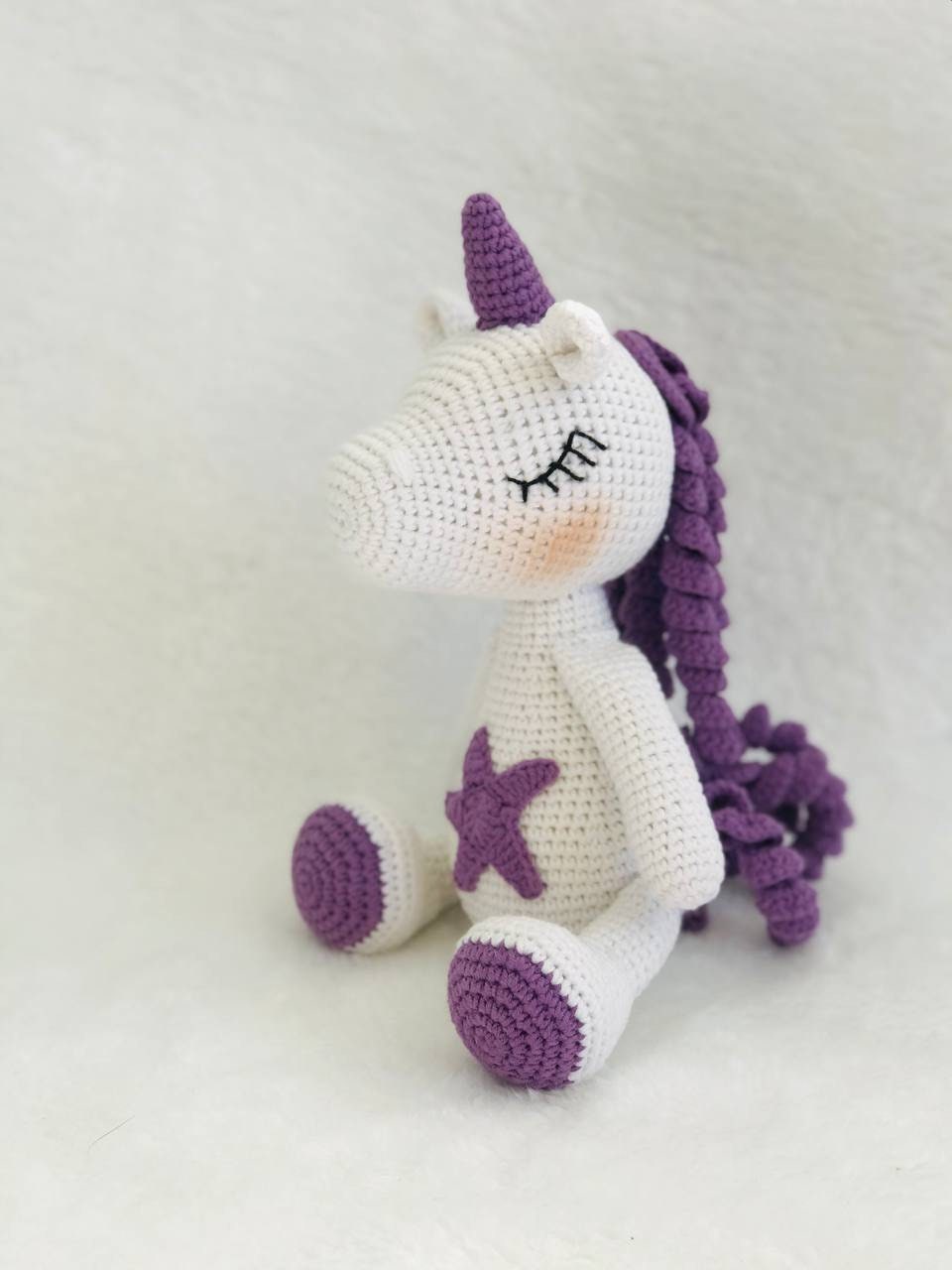 A handmade crochet unicorn plushie in pink and purple, featuring a unique design with a rainbow colored mane and tail. perfect for nursery decor or playtime, and soft and cuddly to the touch. The unicorn doll also has a sparkly horn and pastel colors