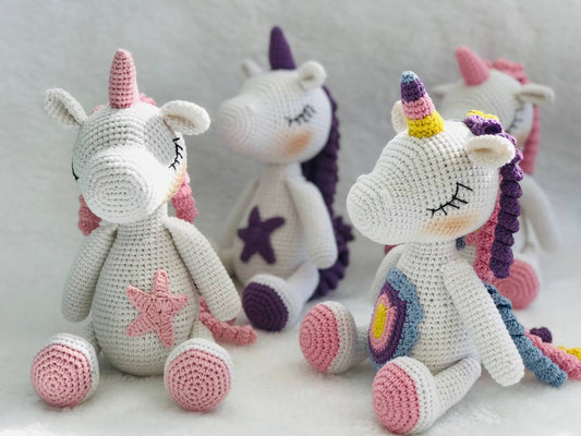 A handmade crochet unicorn plushie in pink and purple, featuring a unique design with a rainbow colored mane and tail. perfect for nursery decor or playtime, and soft and cuddly to the touch. The unicorn doll also has a sparkly horn and pastel colors