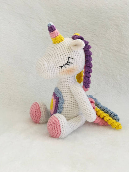 A handmade crochet unicorn plushie in pink and purple, featuring a unique design with a rainbow colored mane and tail. perfect for nursery decor or playtime, and soft and cuddly to the touch. The unicorn doll also has a sparkly horn and pastel colors